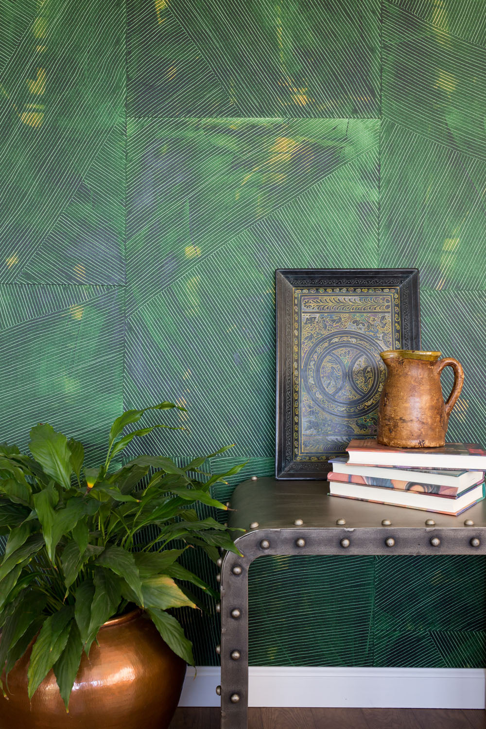 A large plant and an end table stand in front of a wall covered in multi-directional combed wallpaper in mottled green.