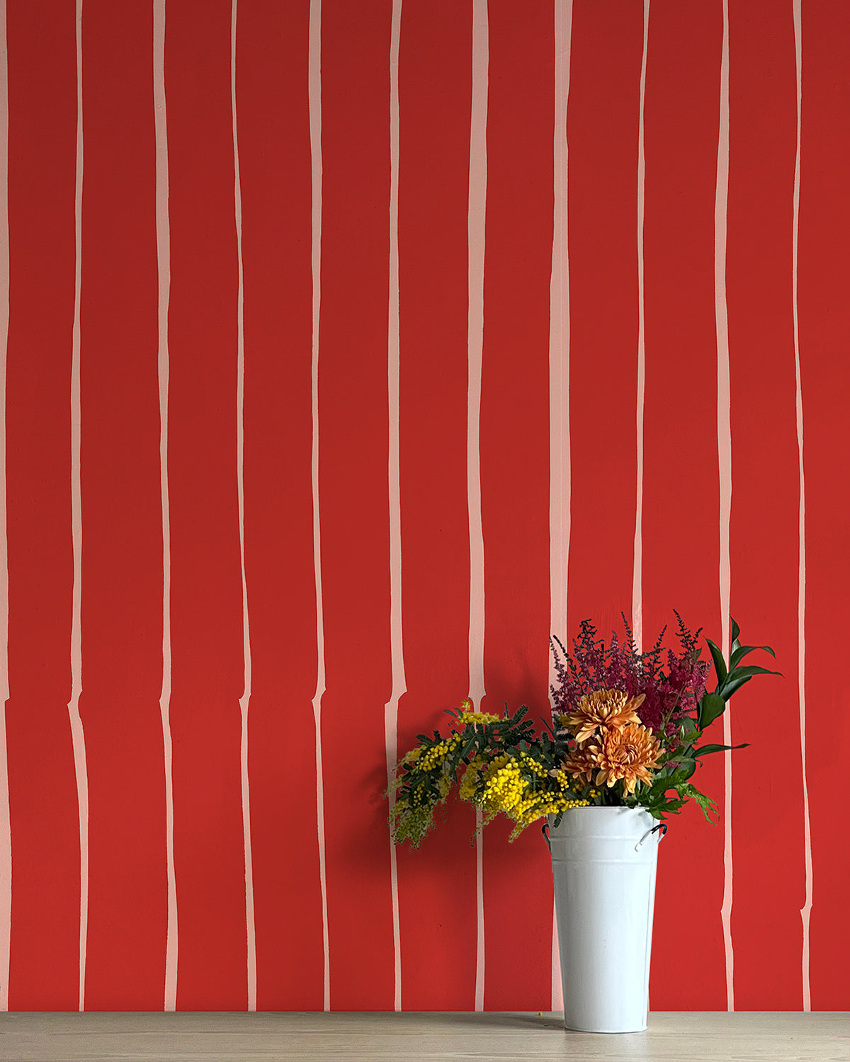 A vase of flowers stands in front of a wall covered in a playful irregular stripe print in cream on a red field.