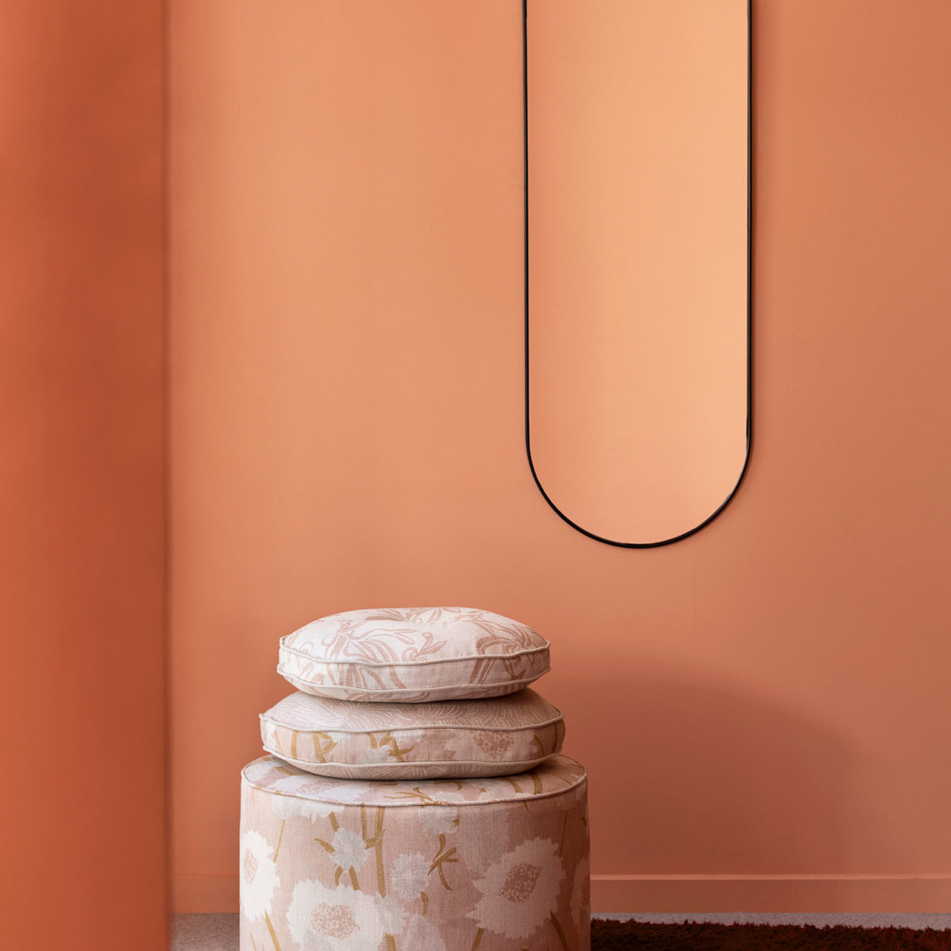 A pouf and stack of pillows, all in a playful floral print in light pink and tan, stand in front of a painted orange wall.