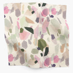 Square fabric swatch in an abstract shape print in shades of pink, purple and green on a cream field.