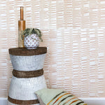 Pillows and an end table stand in front of a wall papered in an undulating ribbon pattern in peach.