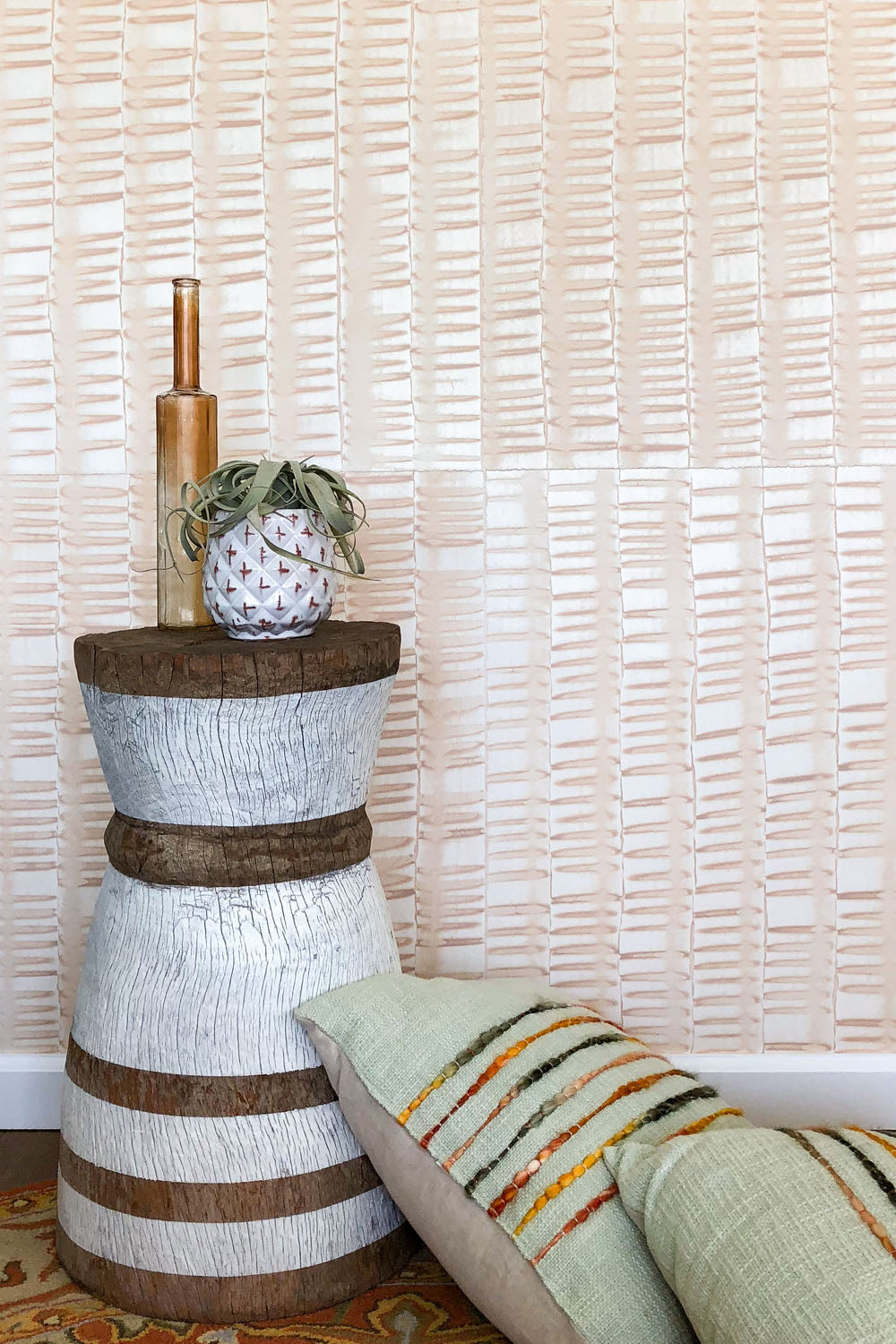 Pillows and an end table stand in front of a wall papered in an undulating ribbon pattern in peach.