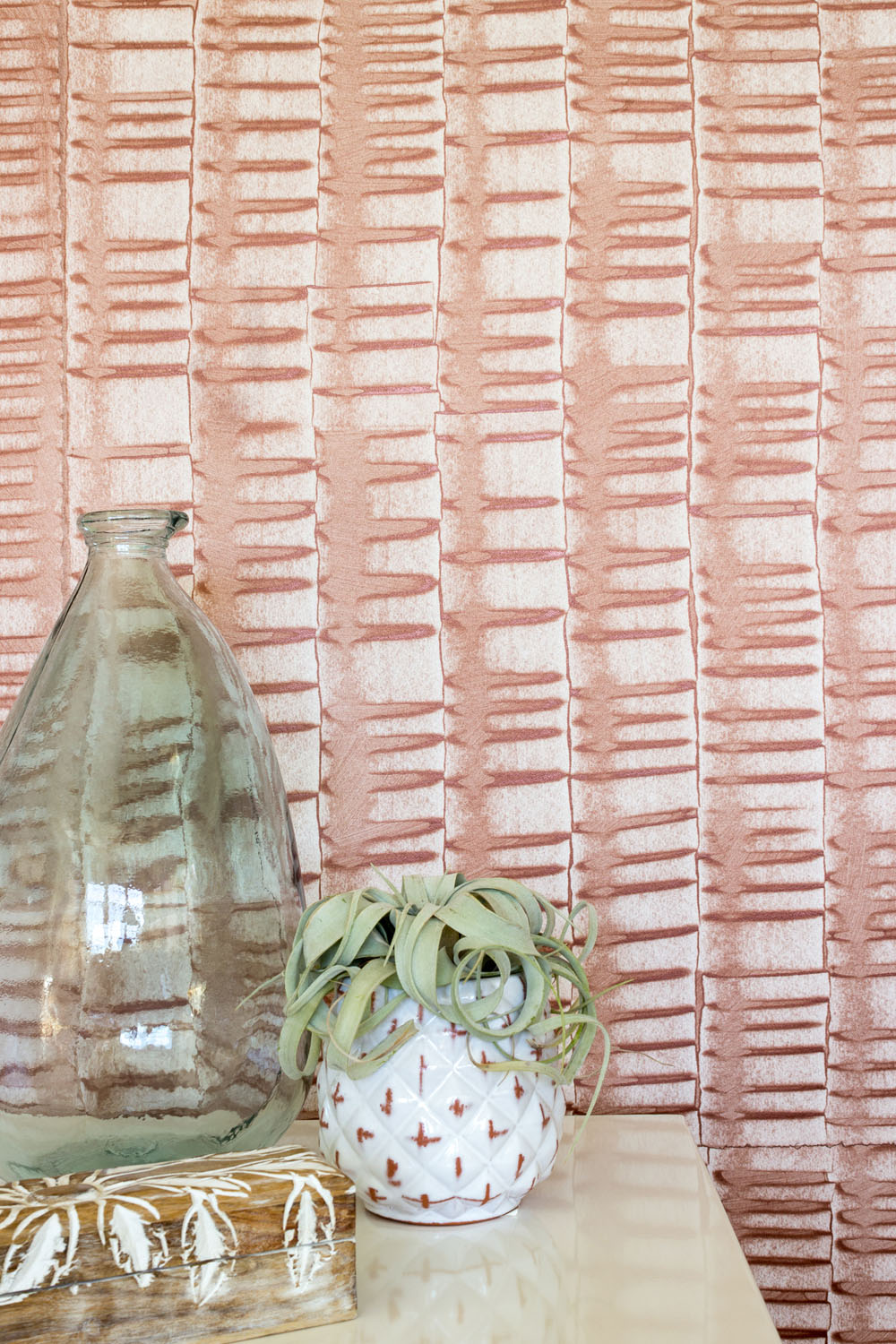 A vase and potted plant stand in front of a wall papered in an undulating ribbon pattern in copper.