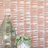 A vase and potted plant stand in front of a wall papered in an undulating ribbon pattern in copper.