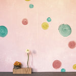 A jar of branches stands in front of a wall papered in a paint splatter print in multicolor pastels on a coral field.