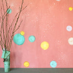A table with knick knacks stands in front of a wall papered in a paint splatter print in pastels on a pink field.