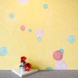 A vase of flowers stands in front of a wall papered in a paint splatter print in multicolor pastels on a yellow field.