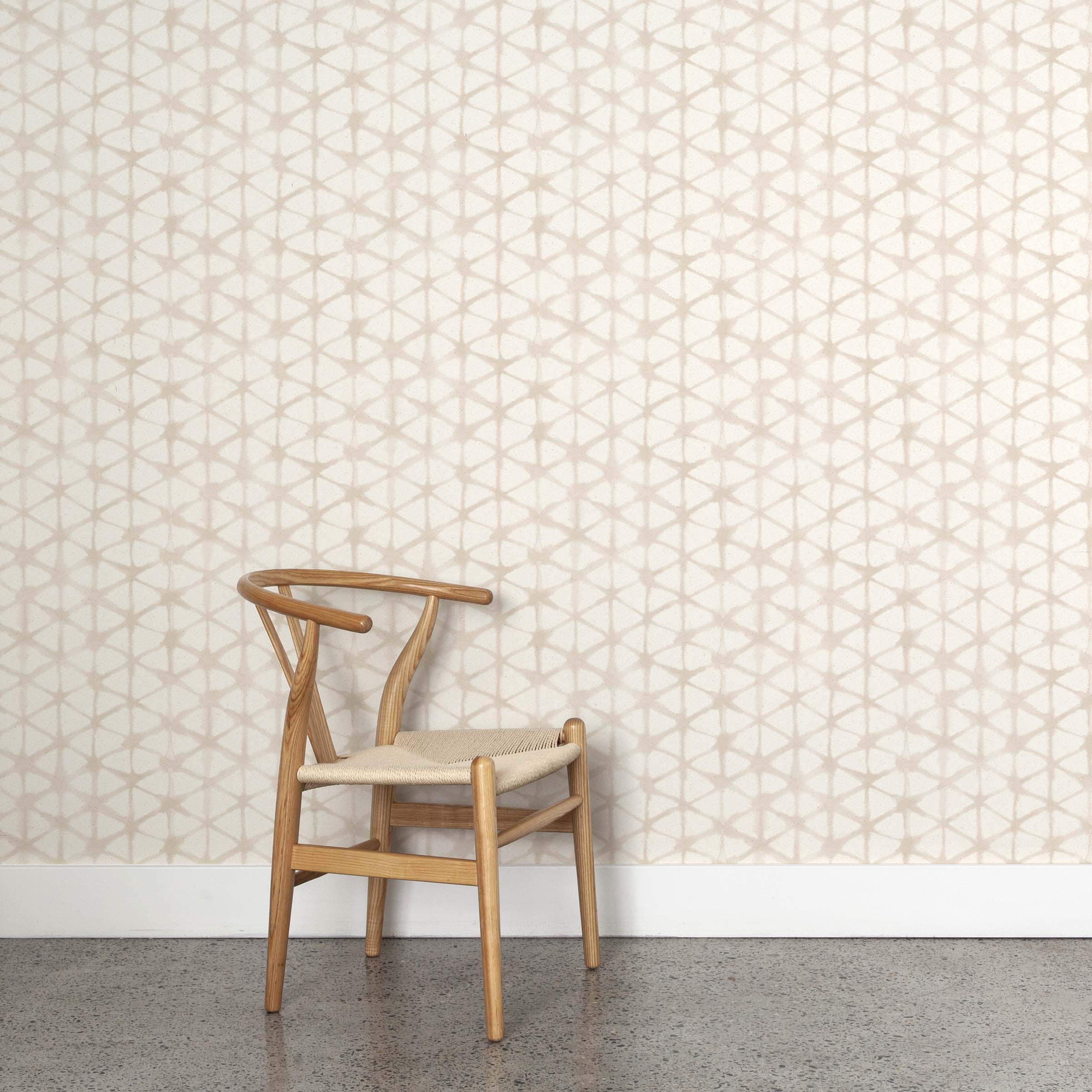 A wooden chair stands in front of a wall papered in a painterly lattice print in tan on a cream field.