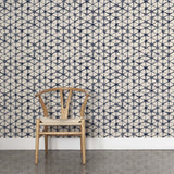 A wooden chair stands in front of a wall papered in a painterly lattice print in indigo on a tan field.