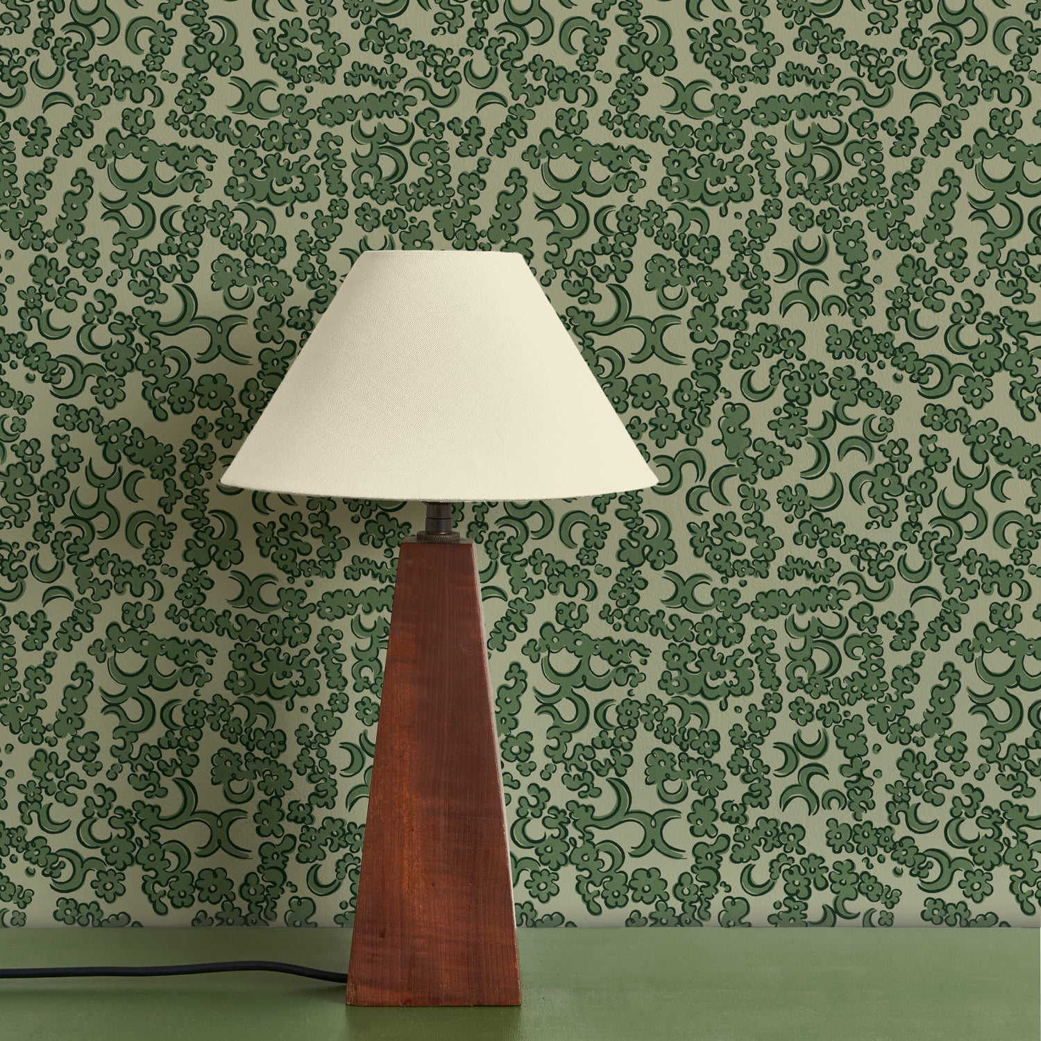 A wooden lamp with ivory shade against botanical lace patterned wallpaper in green. 