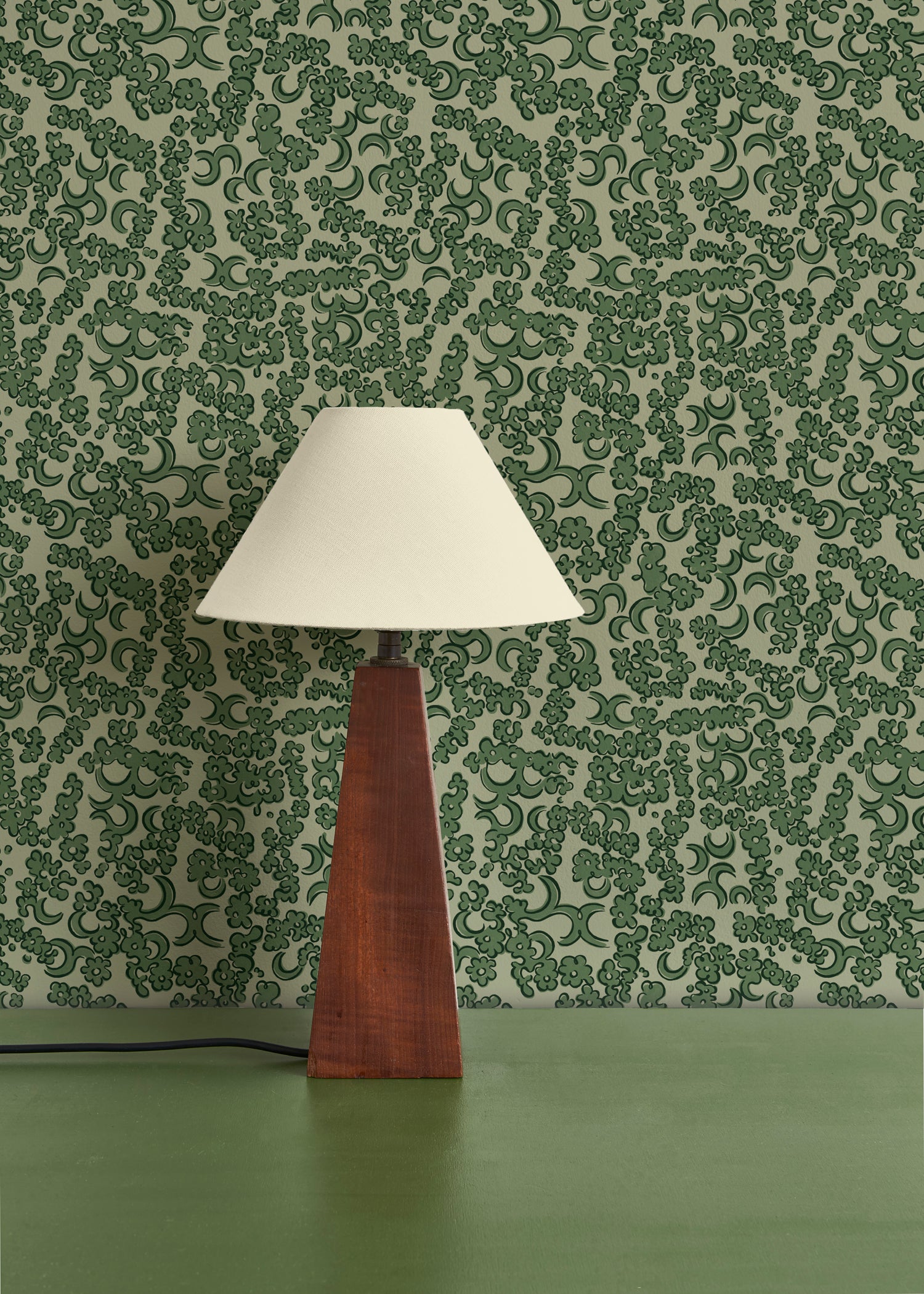 A wooden lamp with ivory shade against botanical lace patterned wallpaper in green. 