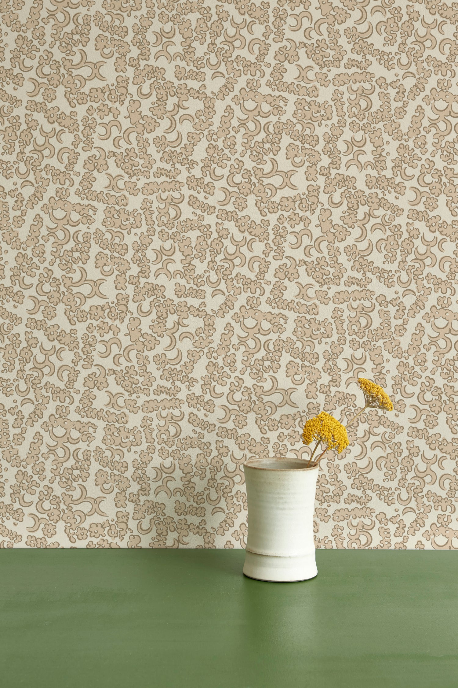 A ceramic vase with yellow wildflowers against wallpaper in a botanical lace pattern in tonal shades of light taupe and ivory.