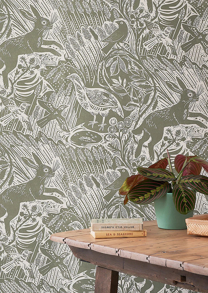 A wooden table with plant and books against a wall papered in a playful rabbit and bird print in white on a moss green field.