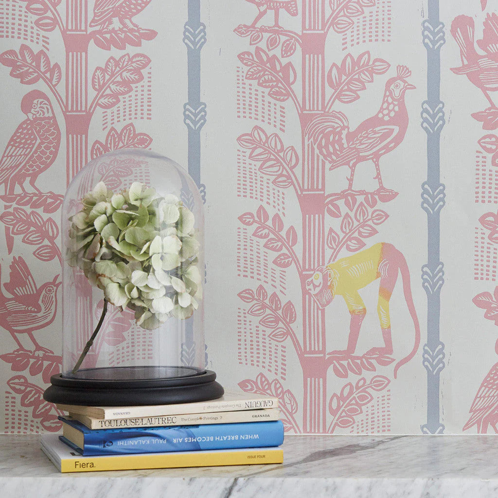 A stack of books stands in front of a wall papered in a linear monkey, bird and leaf print in pink, blue, yellow and cream.