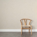 A wooden chair stands in front of a wall papered in a repeating leaf print in white on a cream field.