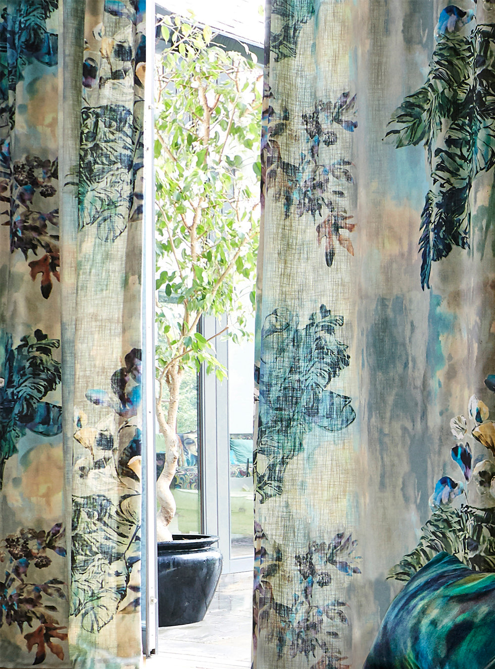 A large potted tree stands behind long curtains in a painterly botanical print.