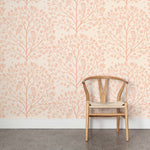 A wooden chair stands in front of a wall papered in a repeating owl and tree print in coral on a cream field.