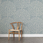 A wooden chair stands in front of a wall papered in a repeating owl and tree print in cream on a light blue field.