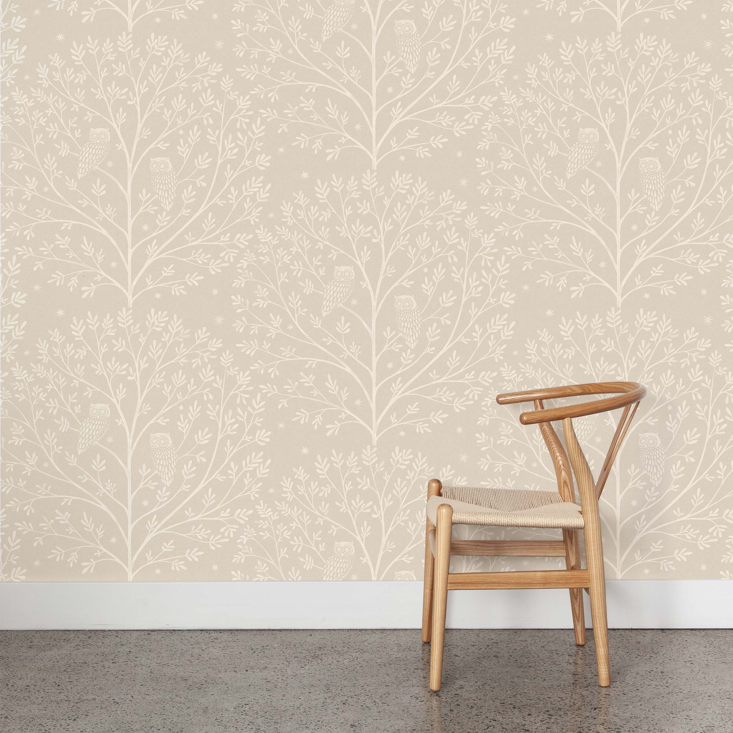 A wooden chair stands in front of a wall papered in a repeating owl and tree print in white on a cream field.