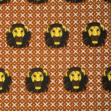 Detail of fabric in a repeating lion face print on a geometric field in shades of red, brown and yellow.