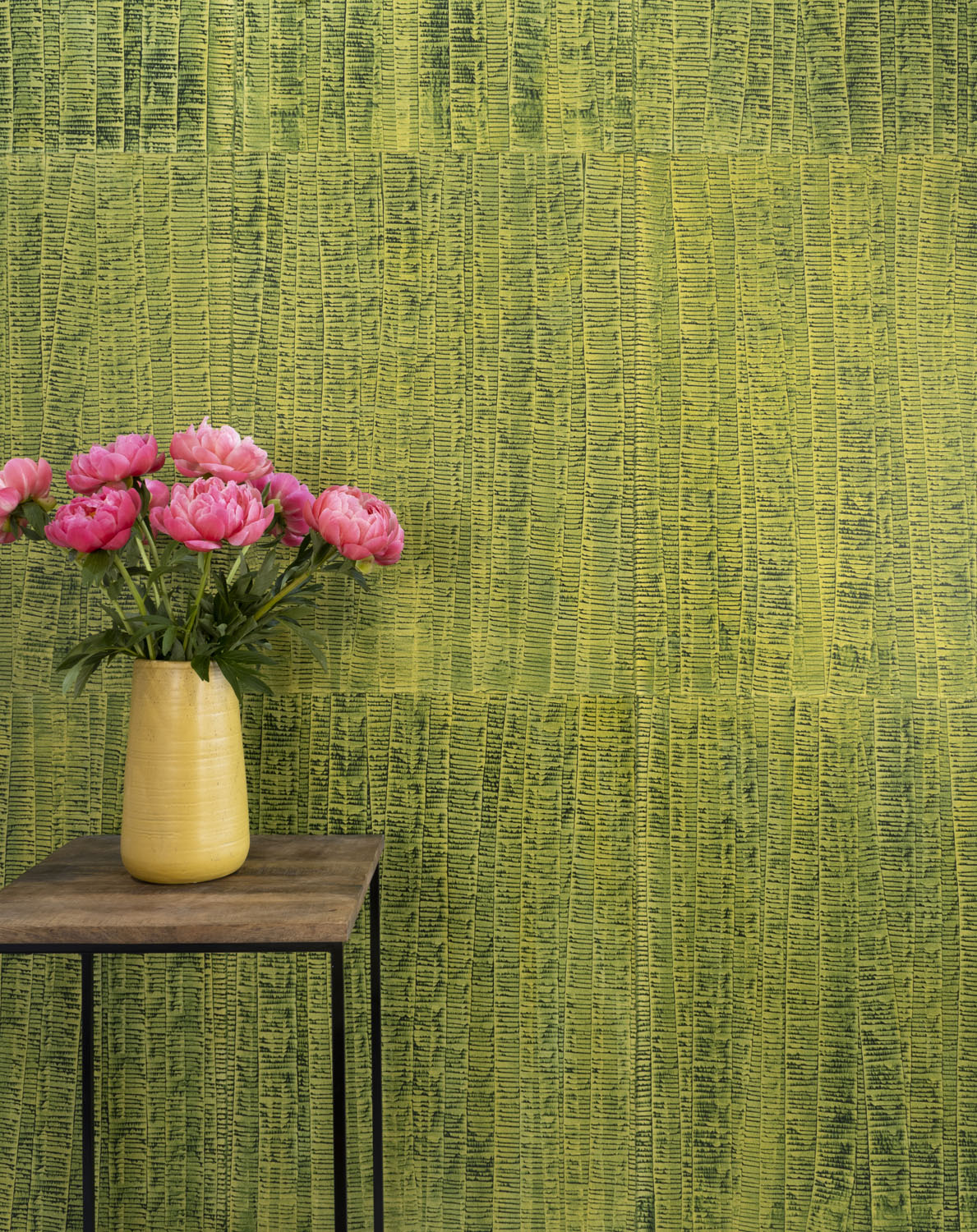 A vase of flowers stands in front of a wall papered in an undulating ribbon pattern in green on a gold field.