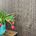 A vase of flowers stands in front of a wall papered in an undulating ribbon pattern in charcoal on a silver field.