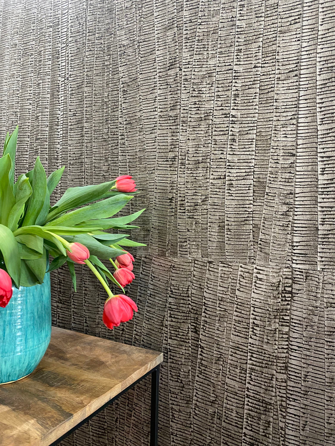 A vase of flowers stands in front of a wall papered in an undulating ribbon pattern in charcoal on a silver field.