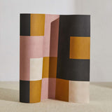 A curved partition stands on the ground, covered in an interlocking square pattern in shades of mustard, pink and gray.