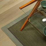 Green rug on a wooden floor with a glass table and wooden chair.