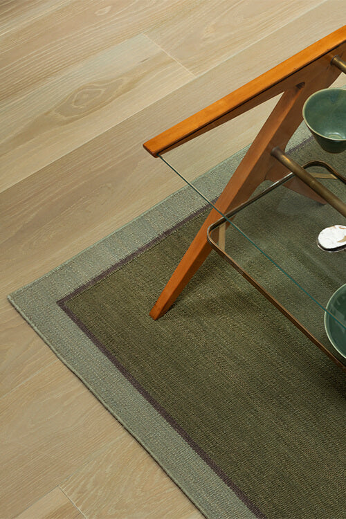 Green rug on a wooden floor with a glass table and wooden chair.