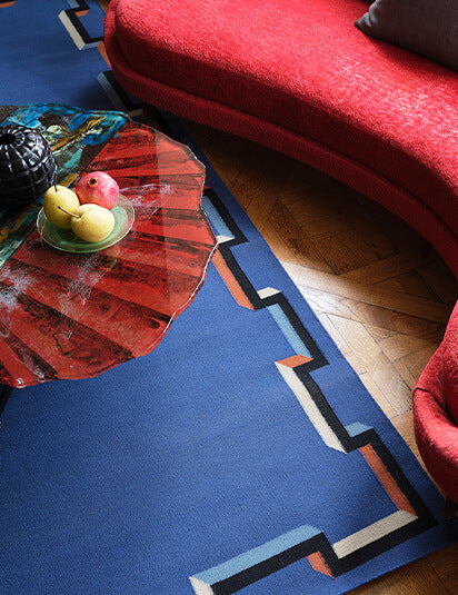 Living room scheme with curved red couch and sculptural coffee table over a a flatwoven rug in a geometric border in blue, black, red and cream.
