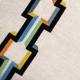 Detail of fabric with a stripe of geometric embroidery in shades of black, orange, yellow and blue on a greige field.