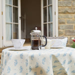 An outdoor table with a french press and mugs, covered in a tablecloth in a floral paisley print.