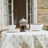 An outdoor table with a french press and mugs, covered in a tablecloth in a floral paisley print.