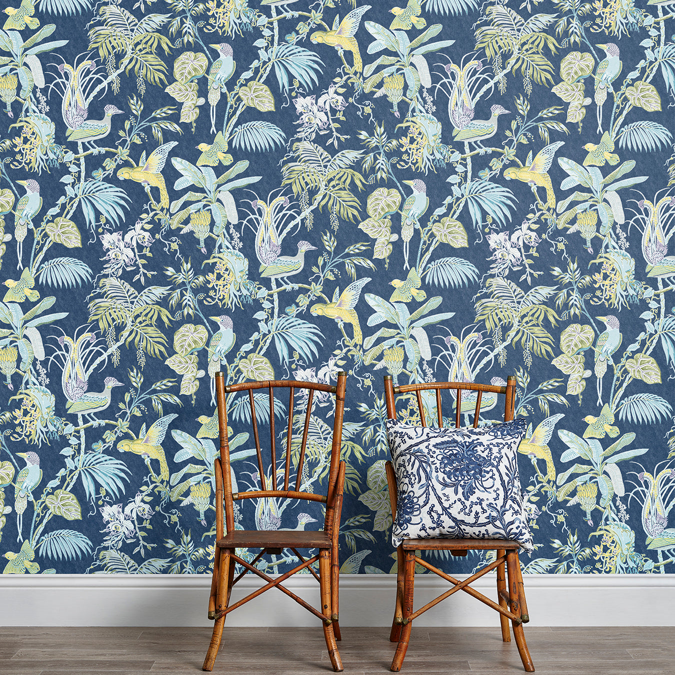 Two wooden chairs stand in front of a wall papered in a dense bird and botanical print in yellow, blue and navy.