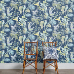 Two wooden chairs stand in front of a wall papered in a dense bird and botanical print in yellow, blue and navy.