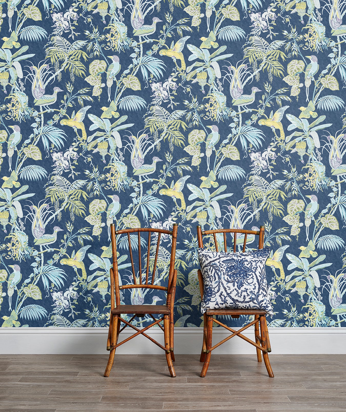 Two wooden chairs stand in front of a wall papered in a dense bird and botanical print in yellow, blue and navy.