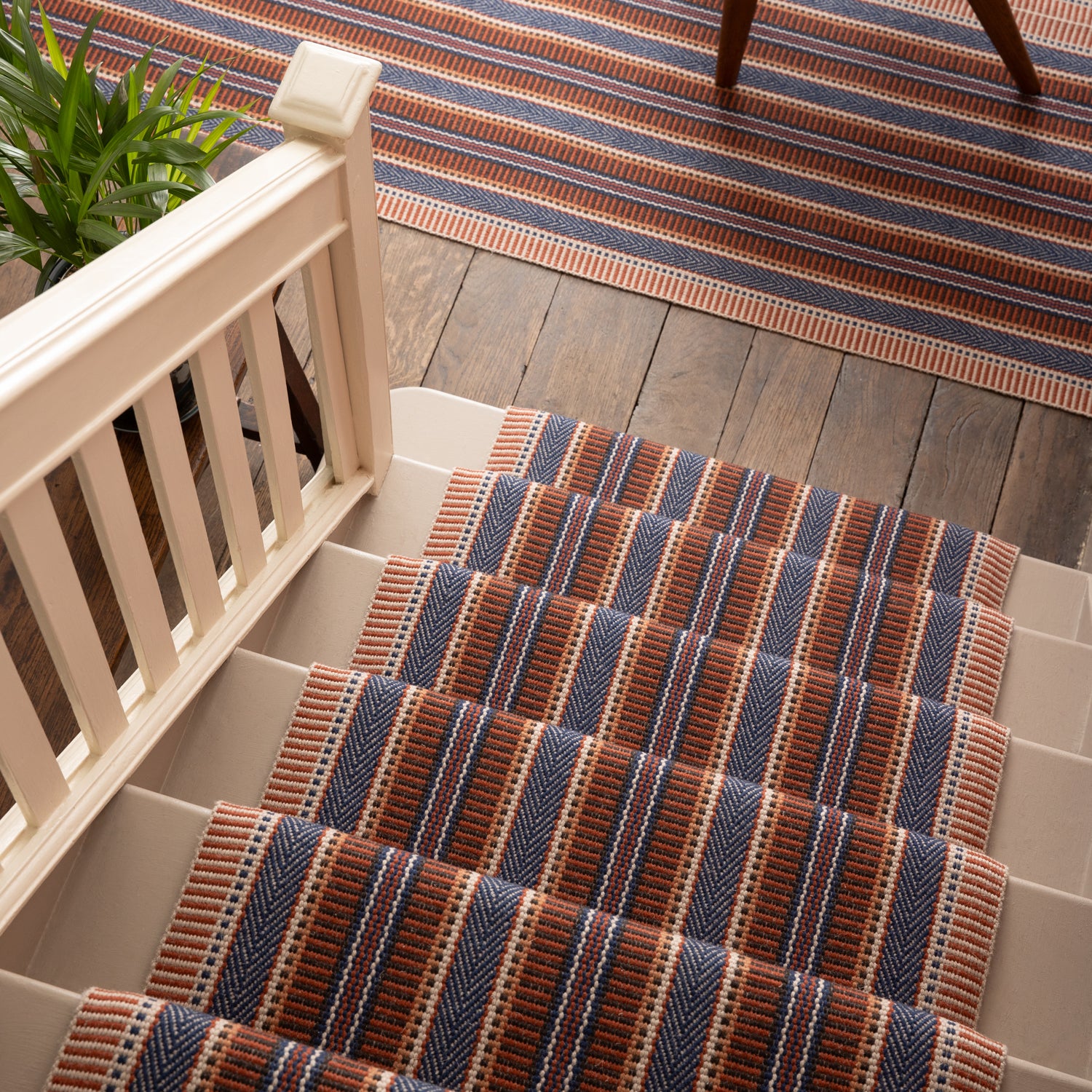 Overhead image of a stair case and landing with a runner and area rug in a complex woven striped made of wool in shades of blue and red. 