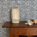 Wooden bookslef with a ceramic vase and conch shells against a blue and cream 'x' patterned wallpaper 