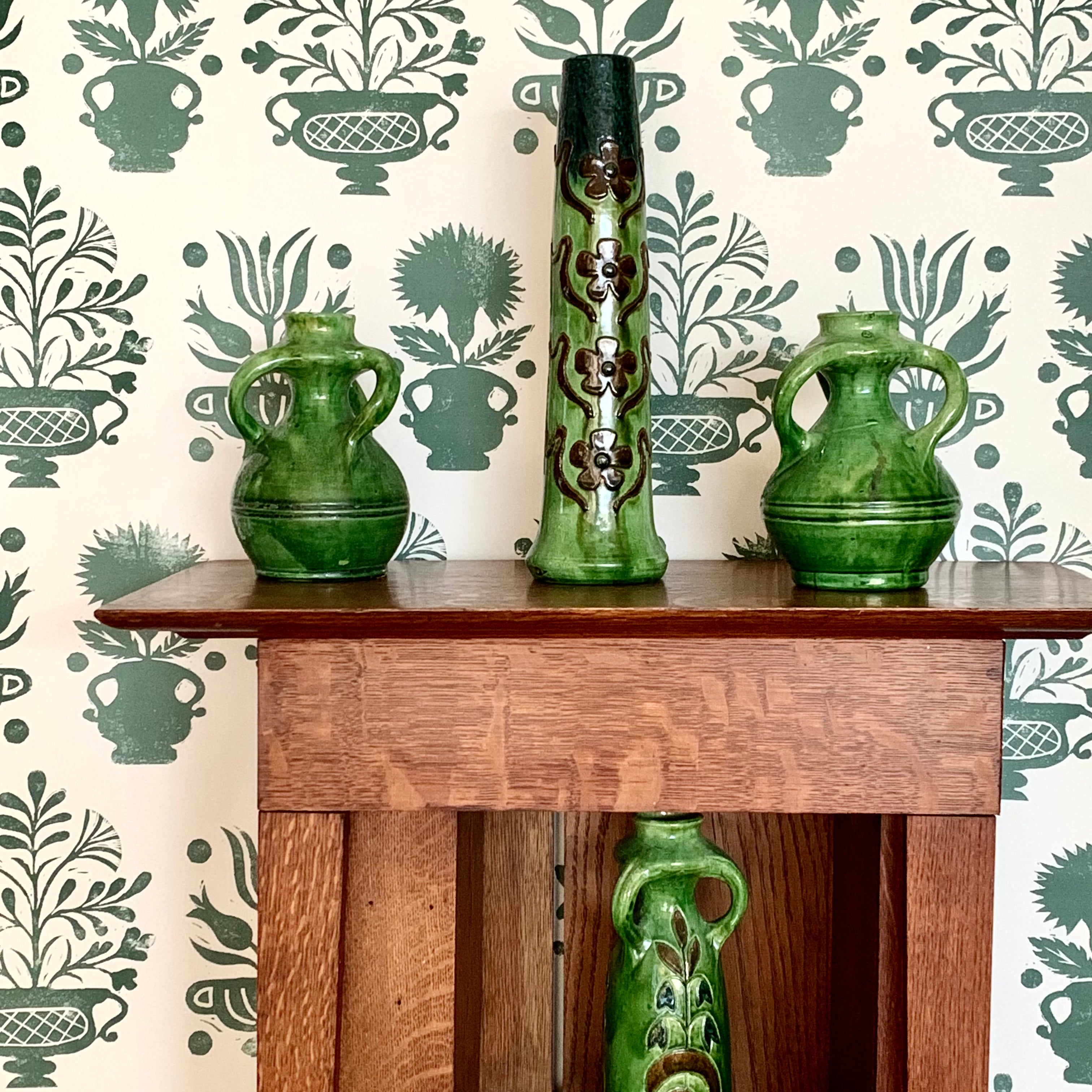 An end table and vases stand in front of a wall papered in a vase and plant print in green on a white field.