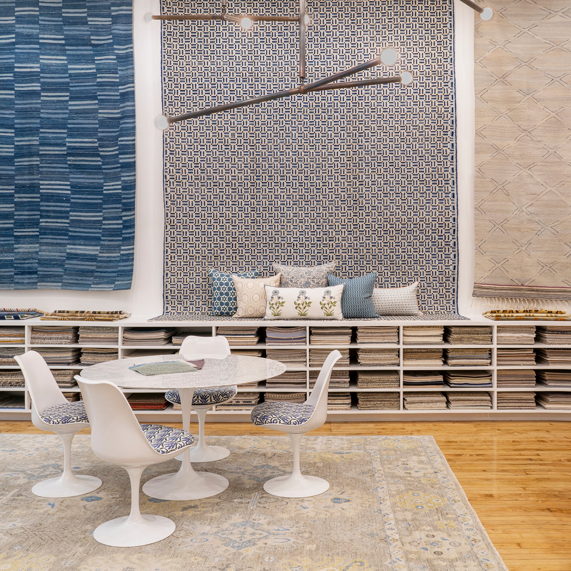 An image of the Studio Four NY Showroom, a white table with four chairs in font of white shelving with rug samples, a display of several pillows against three rugs hanging on the wall. 