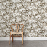 A wooden chair stands in front of a wall papered in a leaf print in shades of brown and tan with a mottled white overlay.
