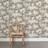 A wooden chair stands in front of a wall papered in a leaf print in shades of brown and tan with a mottled white overlay.