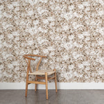A wooden chair stands in front of a wall papered in a leaf print in shades of cream and tan with a mottled white overlay.