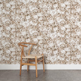 A wooden chair stands in front of a wall papered in a leaf print in shades of cream and tan with a mottled white overlay.