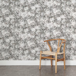A wooden chair stands in front of a wall papered in a leaf print in shades of brown and gray with a mottled white overlay.