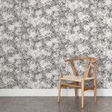 A wooden chair stands in front of a wall papered in a leaf print in shades of brown and gray with a mottled white overlay.
