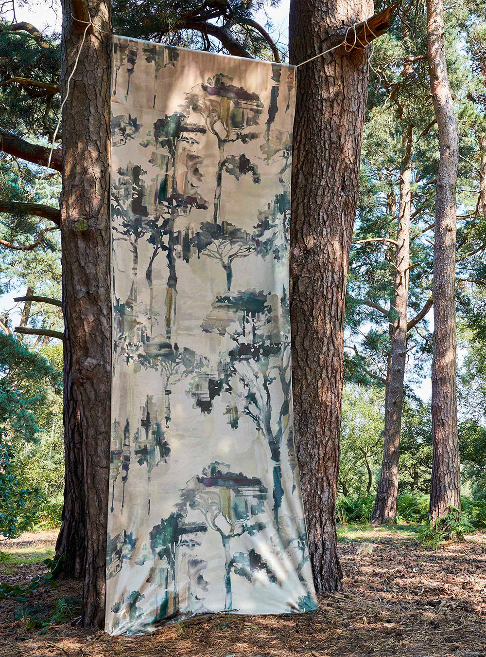 A piece of fabric yardage in a painterly tree print hangs in a forest between two tree trunks.