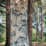 A piece of fabric yardage in a painterly tree print hangs in a forest between two tree trunks.