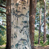 A piece of fabric yardage in a painterly tree print hangs in a forest between two tree trunks.